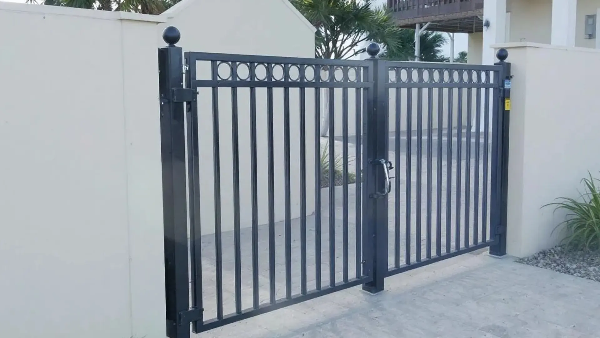 A stylish black aluminum double gate featuring vertical pickets and a decorative ring top design. Mounted between stucco pillars, this gate offers both elegance and durability—perfect for residential or commercial entryways.