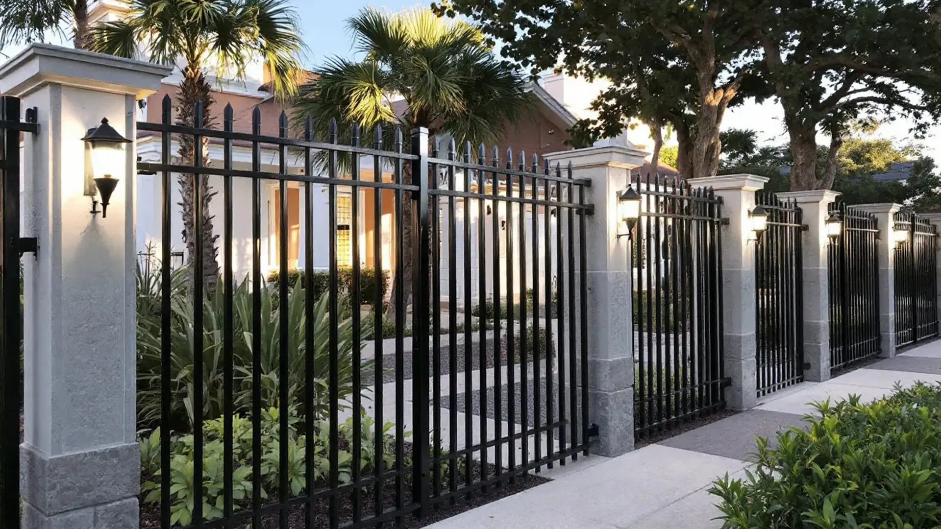 A decorative black aluminum fence with spear-top pickets is framed by tall stone columns fitted with lantern-style lights. The fence encloses a landscaped property with palm trees and lush greenery, creating a sophisticated and secure perimeter around a modern building.