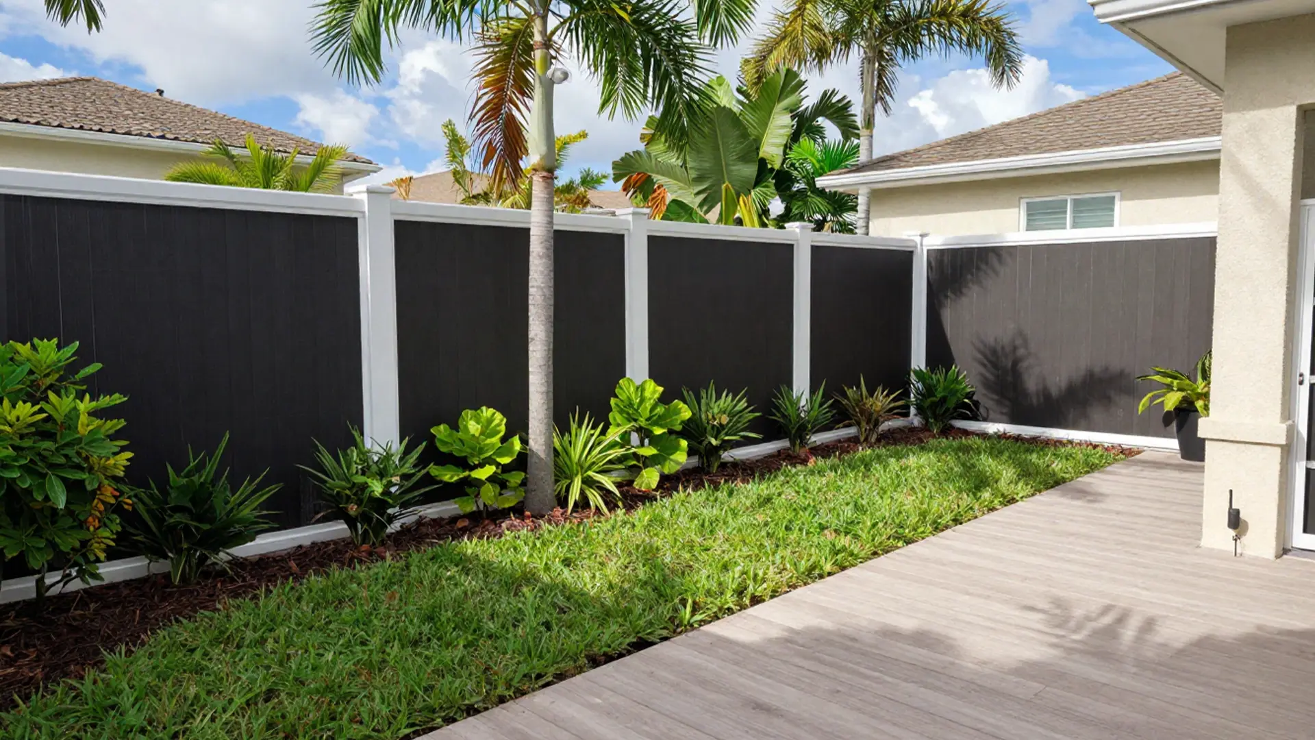 A backyard features a tall black privacy fence with white posts running along the property line. In front of the fence, a landscaped mulch bed contains tropical plants, small shrubs, and palm trees. A strip of green grass borders the planting area, and a light-colored concrete patio extends along the side of a home. Neighboring houses and palm trees are visible beyond the fence under a bright daytime sky.