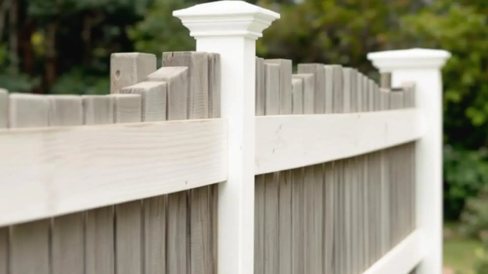 Close-up view of a composite privacy fence with vertical planks in a muted beige tone, topped with white decorative crown-style post caps, set against a backdrop of blurred greenery.