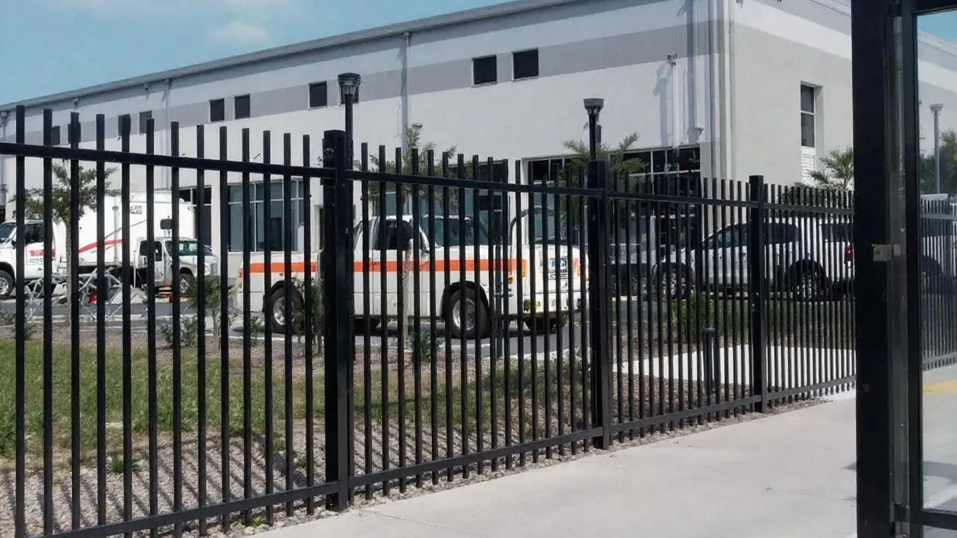 A tall black aluminum bar fence encloses a commercial property with vehicles parked inside, including a white truck with an orange stripe and other cars. The fence stands in front of a large gray-and-white industrial building with windows and exterior lighting.