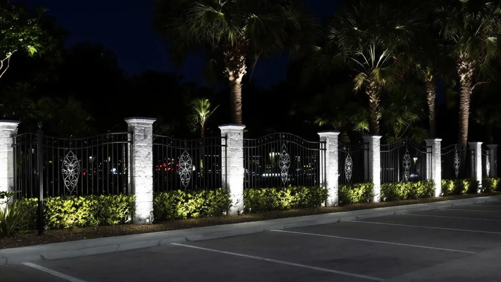 An ornate black wrought iron fence with decorative scrollwork panels is supported by tall stone columns, illuminated by ground lights at night. The lighting highlights the columns and neatly trimmed hedges along the base, while palm trees rise above the fence, creating a dramatic and elegant nighttime setting. The foreground features an empty paved parking area, enhancing the sense of order and security.
