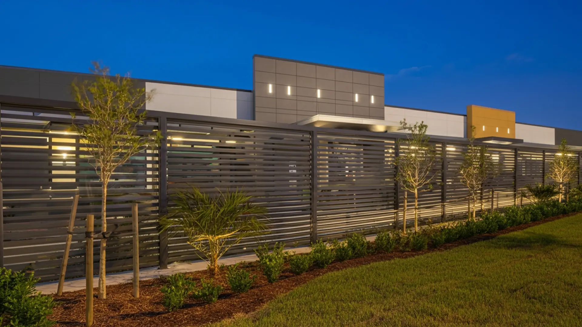 A sleek horizontal metal slat fence lines the front of a modern commercial building, illuminated by warm exterior lights at dusk. Young trees and neatly trimmed shrubs are planted along the fence, with a manicured lawn in the foreground and the contemporary building with geometric panels in the background.