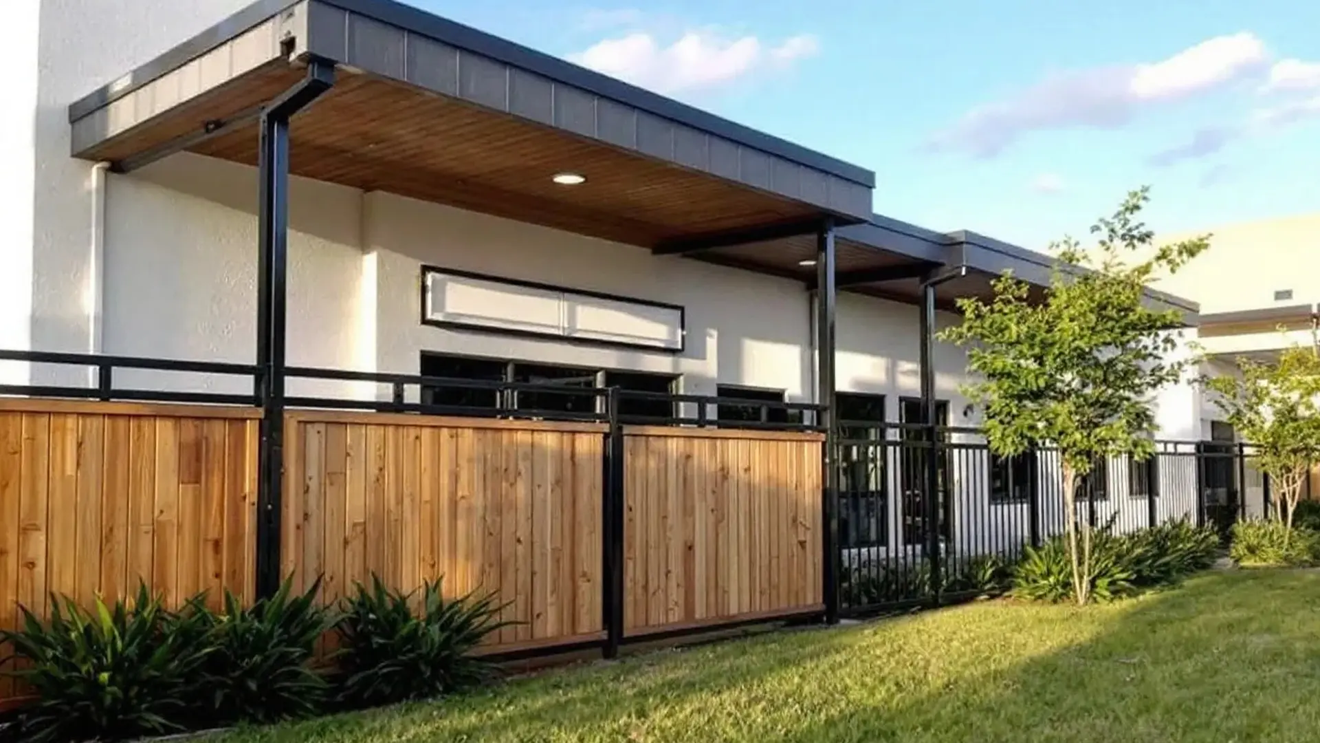 A contemporary building with a flat roof and wood-paneled ceiling overhang. In front, a privacy fence combines vertical natural wood planks framed with black metal posts and railings. Lush green landscaping with shrubs and young trees lines the base of the fence, set against a grassy lawn under a bright sky.