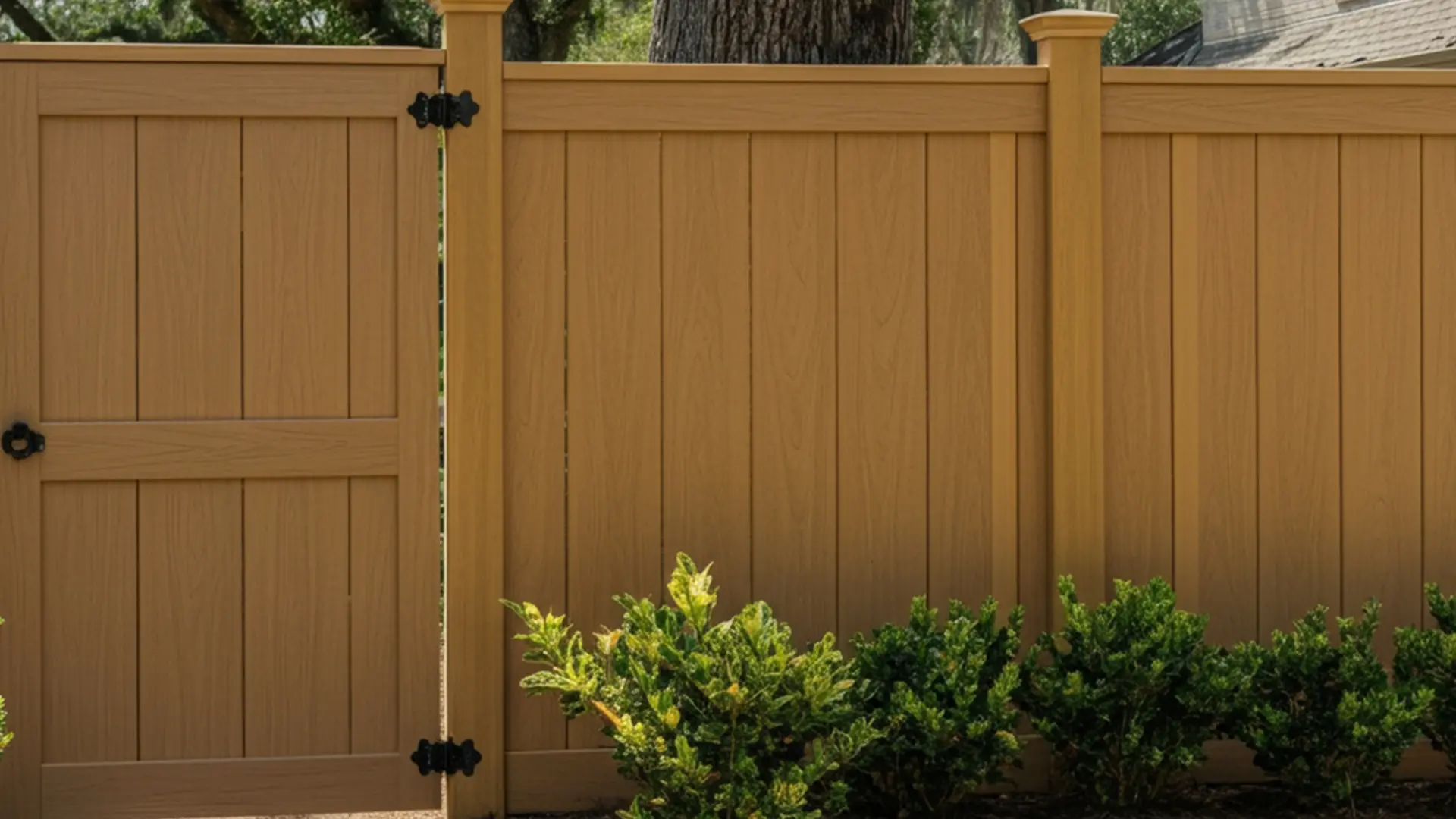 Solid composite fence in a warm tan color featuring vertical planks, decorative crown post caps, and a matching gate with black metal hinges and latch, bordered by small green shrubs.