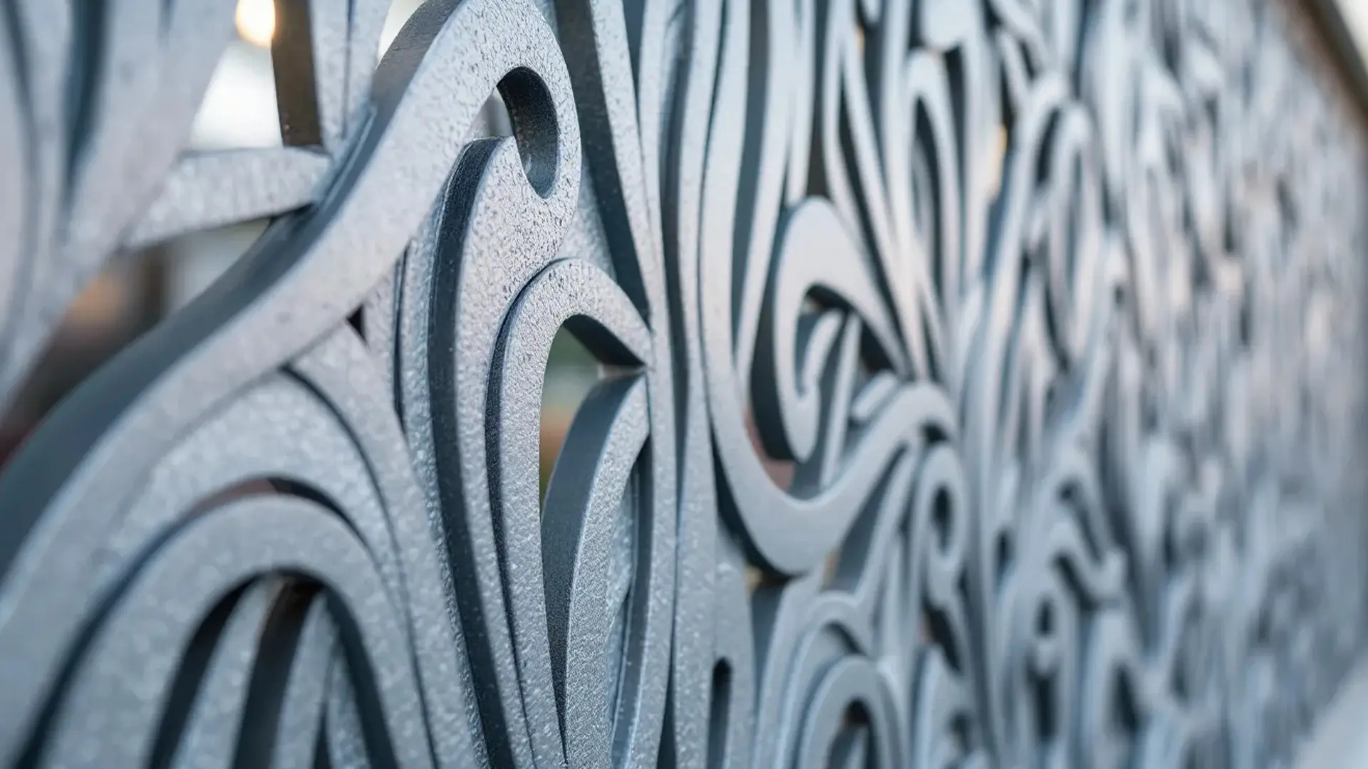 A close-up view of a decorative metal privacy screen featuring an abstract swirling cut-out pattern. The textured gray surface catches the light, with the background softly blurred to emphasize the intricate design in the foreground.